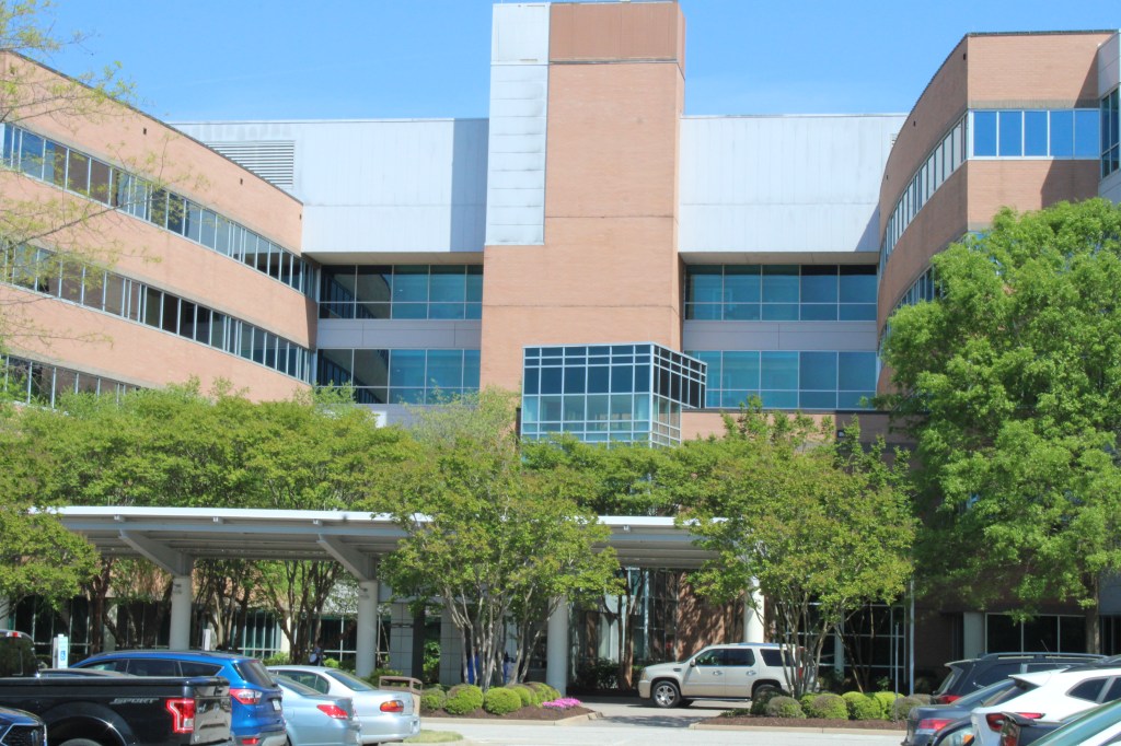A hospital on a sunny day with a full parking lot and trees in between the parking lot.