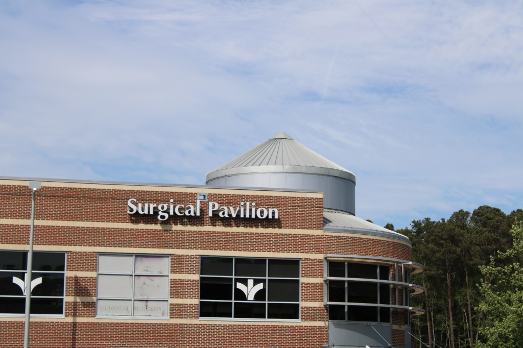 The outside of a surgical pavilion during a bright and sunny day, next to trees.